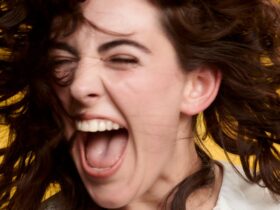 A girl with brown hair shakes her head while laughing against a yellow background.