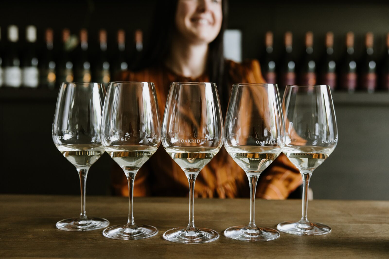 5 wine glasses, each with a tasting of chardonnay. A smiling woman can be seen in the background.