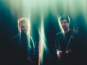 2 short haired males holding string instruments in spotlight of fragmented light