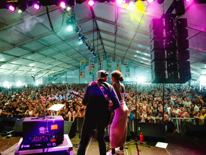 A woman sings and a man plays guitar, with hundreds of people inside a tent