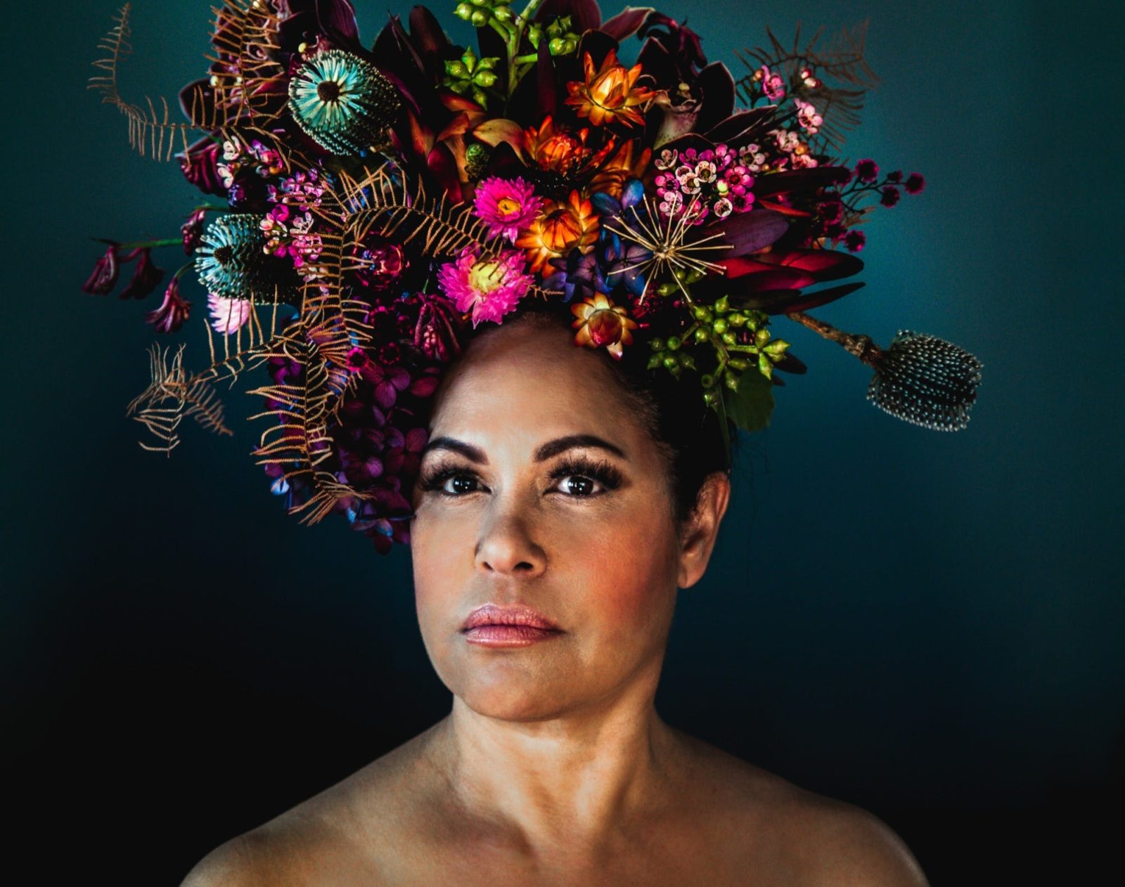 Christine Anu, shoulders up, is looking at the camera wearing a large colourful flower headpiece