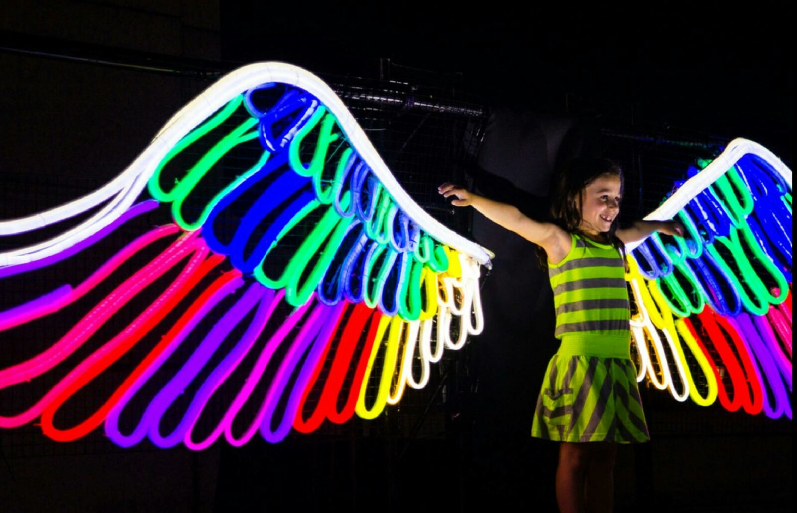 A young girl standing in front of colourful neon wings at night time