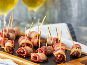 A small bite sized date wrapped in bacon held together with pick, served on wooden board.