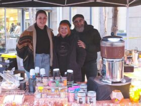 A young woman, man and woman stand behind their market table, displaying hot chocolate to purchase