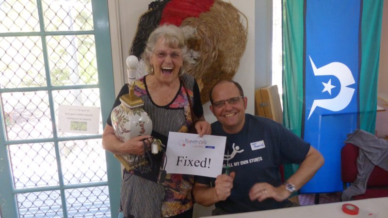 Smiling person holding a lamp and a sign saying 'Fixed', beside the fixer who fixed it