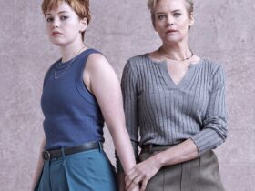 Two women standing side by side, one gently holding the other's hand.