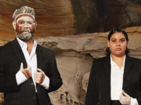 two Indigenous Australians in front of a rock face with painted faces wearing suits
