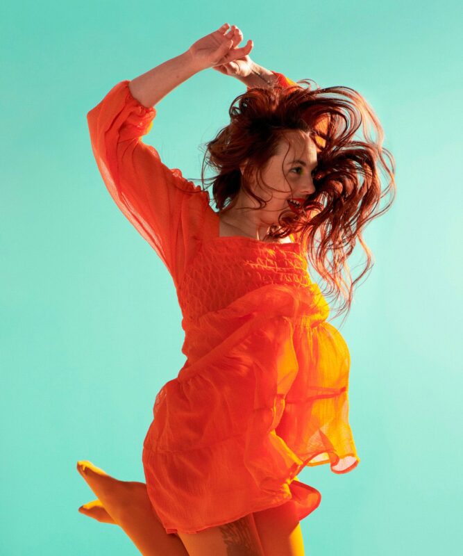 A woman with brown hair wearing an orange dress jumping in the air against a blue background.