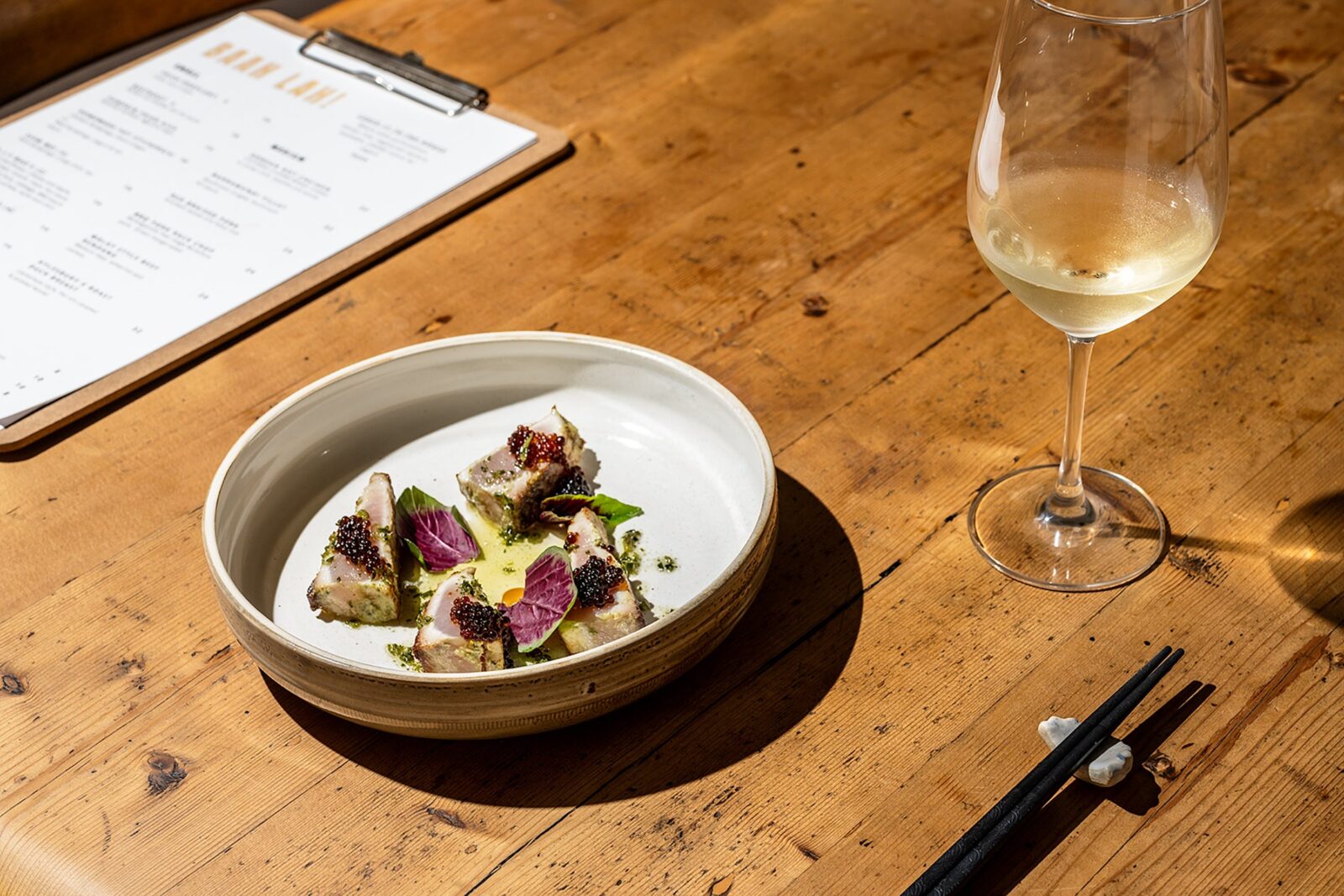 handmade ceramic bowl with seared kingfish pieces inside, a glass of white wine, a menu on table