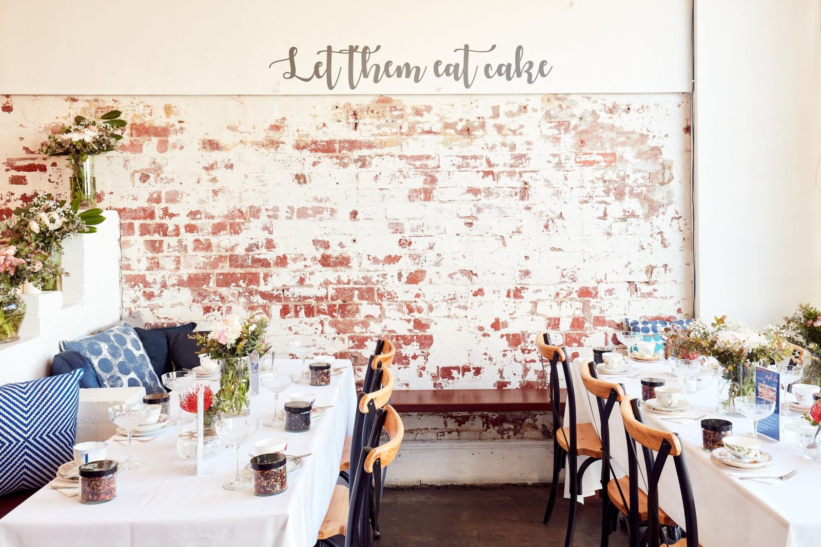 Exposed brick wall, with cursive 'Let them eat cake' written above. Two tables set for high tea.