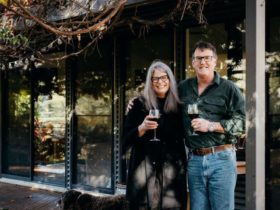 Owners Julie & Dean standing together under verandah with a glass of red wine in hand.