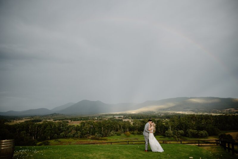 A couple embracing at The Riverstone Estate with a rainbow in the background.