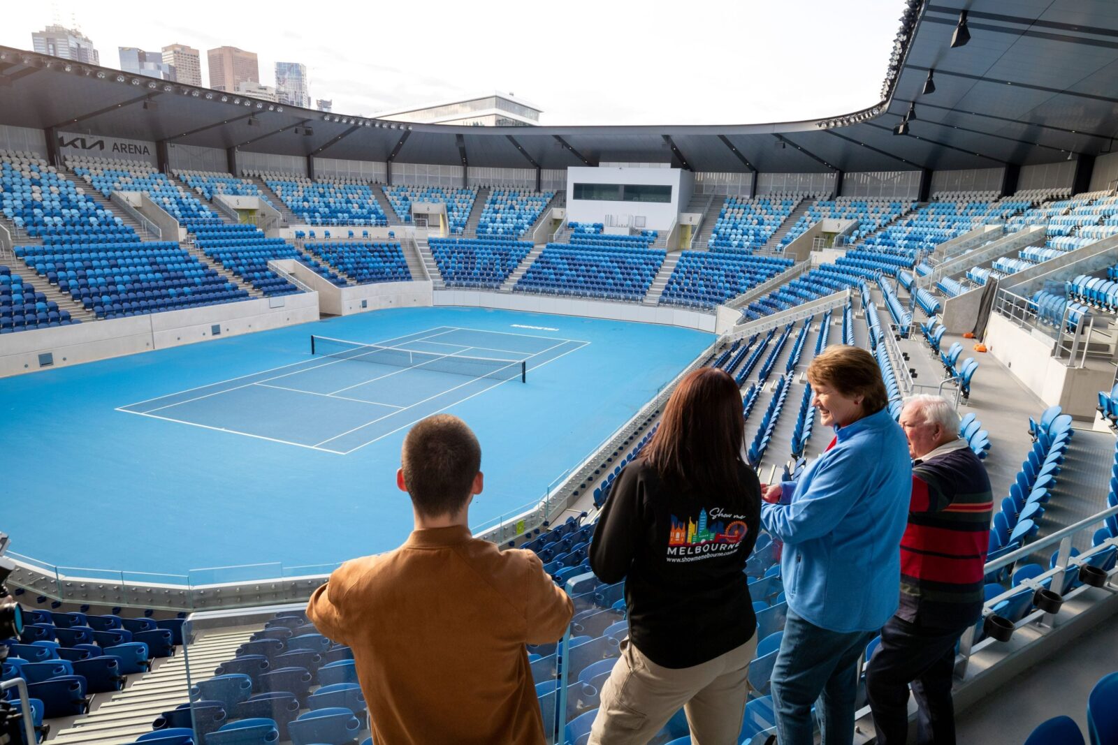 A group of four people are looking at a blue tennis court from the stands. One is a tour guide.