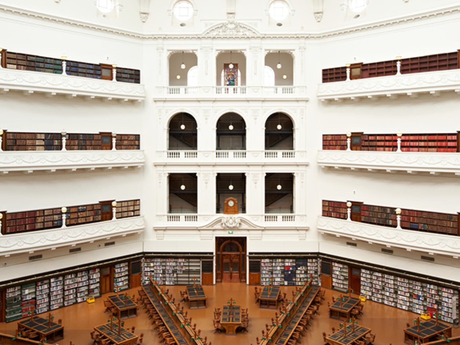 A large, bright white room. Up the top of the image are stacks of bookshelves. Below are desks.