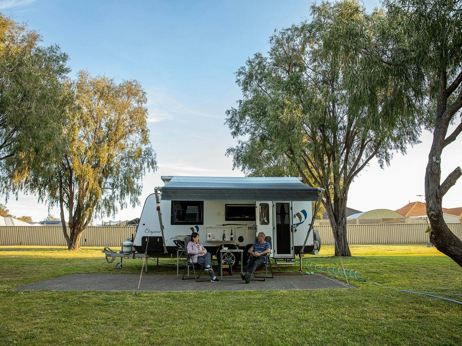 A couple sit outside their caravan surrounded by green grass and peppermint trees