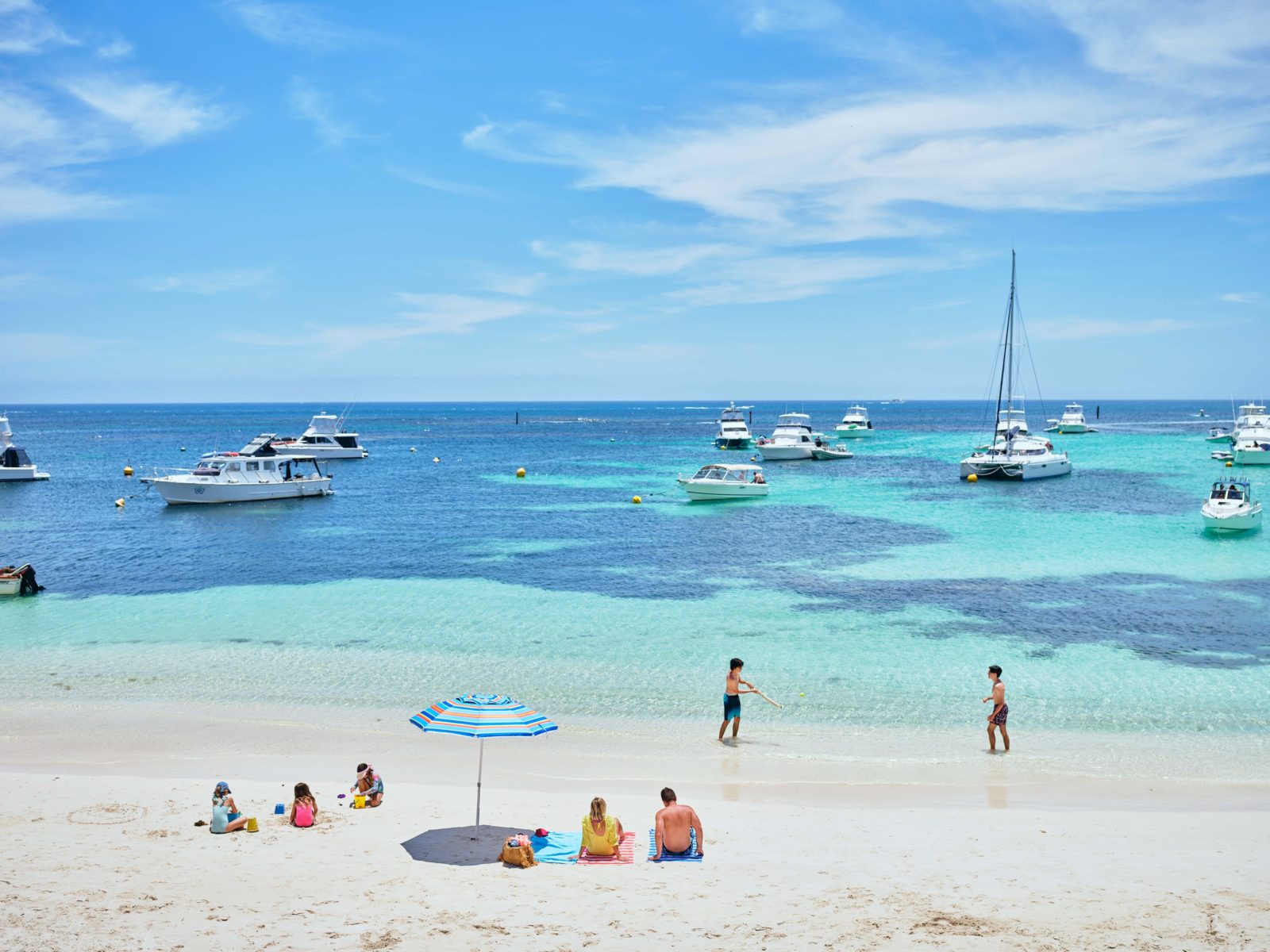 People sitting on a white sandy beach, looking at boats on the water.