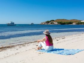 Woman sitting on a towel on a white sandy beach looking out over blue water.
