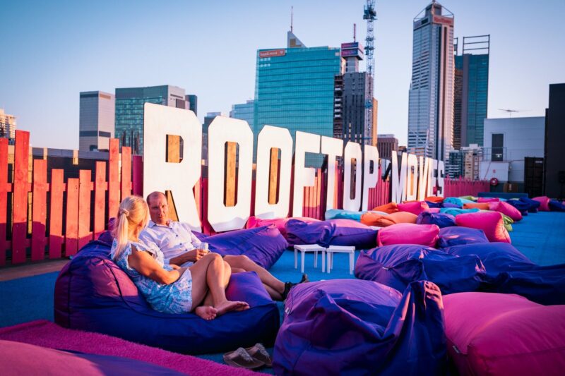Couple sit on beanbags beside the Rooftop Movies sing with the Perth city skyline in the background.