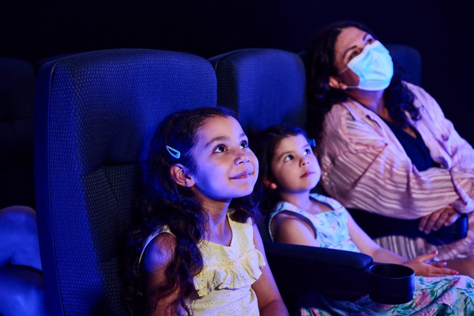 Two small girls and their mother watch the cinema experience, blue light filling the room