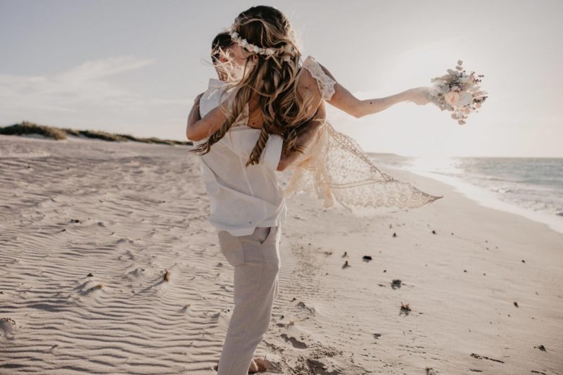 Groom lifting his bride into his arms, overlooking a sunset beach.