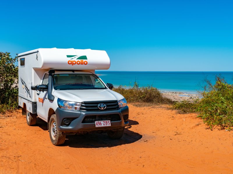 An Apollo Adventure Camper is parked on red dirt near the ocean